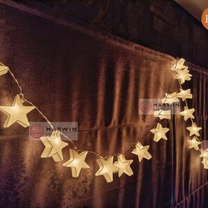 May include: A string of white star-shaped lights with a warm glow. The lights are strung across a brown fabric background. The text "Marwin Craft" is visible on the background.