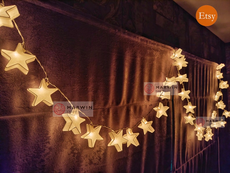 May include: A string of white star-shaped lights with a warm glow. The lights are strung across a brown fabric background. The text "Marwin Craft" is visible on the background.
