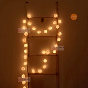 May include: A string of white cotton ball lights draped over a wooden ladder. The lights are lit and glowing a warm yellow. The image includes the text "Etsy" and "Marwin Craft".