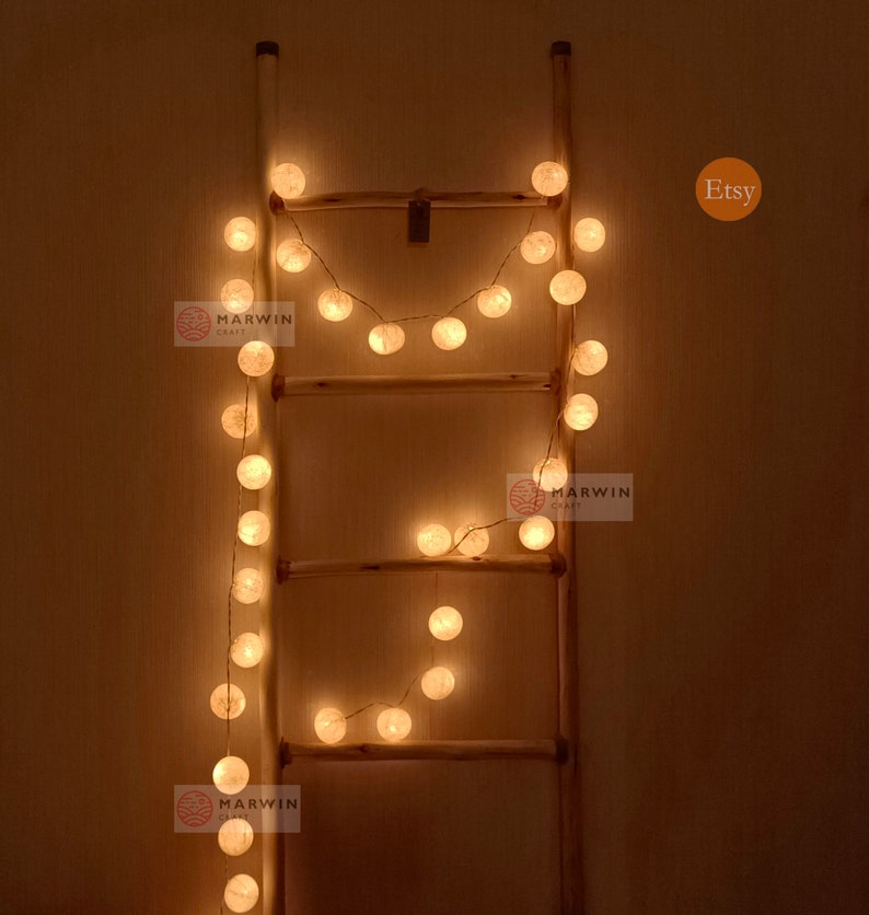 May include: A string of white cotton ball lights draped over a wooden ladder. The lights are lit and glowing a warm yellow. The image includes the text "Etsy" and "Marwin Craft".