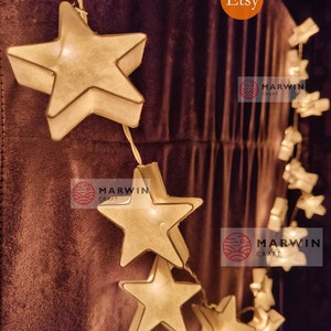 May include: A string of white paper star lights with warm white LED bulbs. The lights are strung on a clear wire and are perfect for adding a touch of whimsy to any room. The lights are hung on a brown fabric background. The image also includes the text "Etsy" and "Marwin Craft".