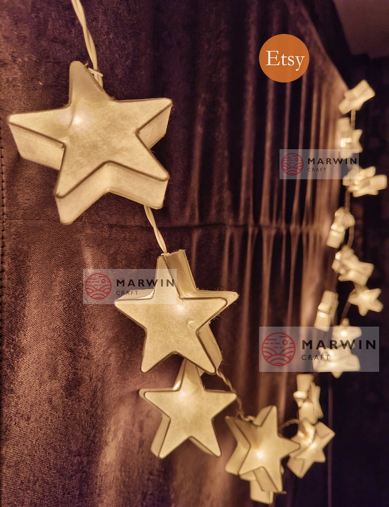 May include: A string of white paper star lights with warm white LED bulbs. The lights are strung on a clear wire and are perfect for adding a touch of whimsy to any room. The lights are hung on a brown fabric background. The image also includes the text "Etsy" and "Marwin Craft".