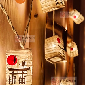 K&ouml;nnte beinhalten: Lichterkette aus wei&szlig;en Papierlaternen mit roten Kreisen und japanischen Schriftzeichen. Die Laternen sind mit warmwei&szlig;en LED-Lichtern beleuchtet. Die Laternen h&auml;ngen an einer Holzwand.