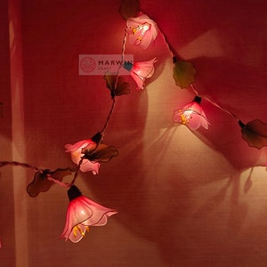 May include: A string of fairy lights with pink flower-shaped bulbs. The lights are strung on a wire with green leaves. The lights are lit and casting a warm glow. The image also includes the text "Etsy" and "Marwin Craft".