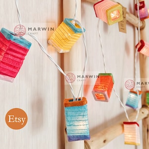 Pode incluir: Cordão de luzes com lanternas de papel coloridas. As lanternas têm formato quadrado e têm um fio branco que as atravessa. As lanternas estão acesas e brilhando. O texto "Marwin Craft" é visível na imagem.