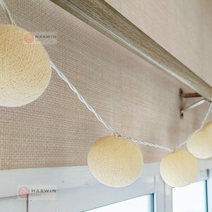 May include: A string of white fairy lights with cream-colored cotton ball bulbs. The lights are strung across a wooden curtain rod and hang down towards a window. The text "MARWIN CRAFT" is visible on the image.