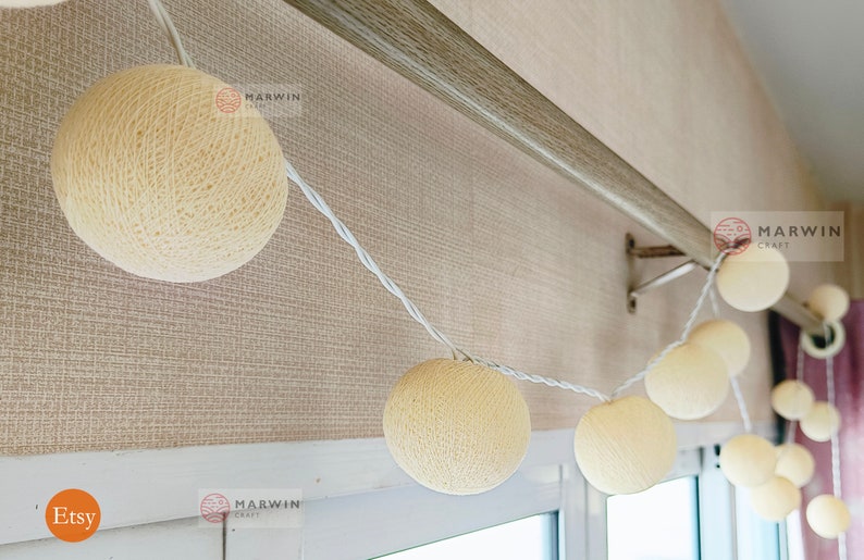 May include: A string of white fairy lights with cream-colored cotton ball bulbs. The lights are strung across a wooden curtain rod and hang down towards a window. The text "MARWIN CRAFT" is visible on the image.