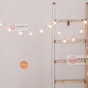 May include: White star-shaped string lights draped over a wooden ladder. The lights are lit and create a warm glow. The ladder is made of natural wood and has a rustic look. The image also includes the text "MARWIN CRAFT" and "Etsy".