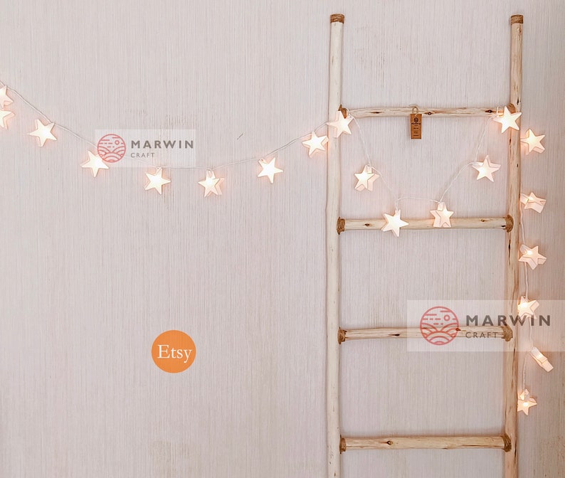 May include: White star-shaped string lights draped over a wooden ladder. The lights are lit and create a warm glow. The ladder is made of natural wood and has a rustic look. The image also includes the text "MARWIN CRAFT" and "Etsy".
