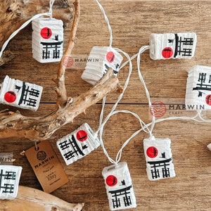 K&ouml;nnte beinhalten: Eine Girlande aus wei&szlig;en Papierlaternen mit roten und schwarzen japanischen Schriftzeichen und einem roten Kreis mit einer Vogelsilhouette. Die Laternen sind an einem wei&szlig;en Kabel befestigt und h&auml;ngen an einem Ast. Die Laternen sind mit den japanischen Schriftzeichen f&uuml;r "Gl&uuml;ck" verziert.