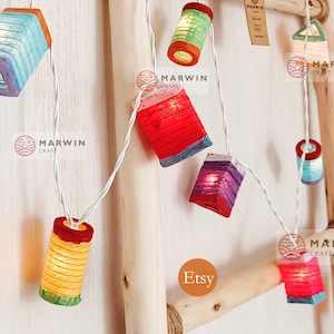 Può includere: Ghirlanda di luci con lanterne di carta colorate in varie forme, tra cui quadrati, rettangoli e cilindri. Le lanterne sono illuminate da luci LED bianco caldo. Le luci sono infilate su un filo bianco e sono appese a una scala di legno. Il testo "Etsy" e "Marwin Craft" è visibile nell'immagine.