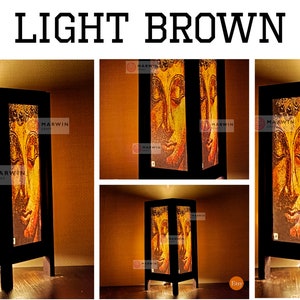 May include: A light brown wooden table lamp with a square base and a rectangular shade. The shade features a printed image of a golden Buddha face. The lamp is lit and casting a warm glow.