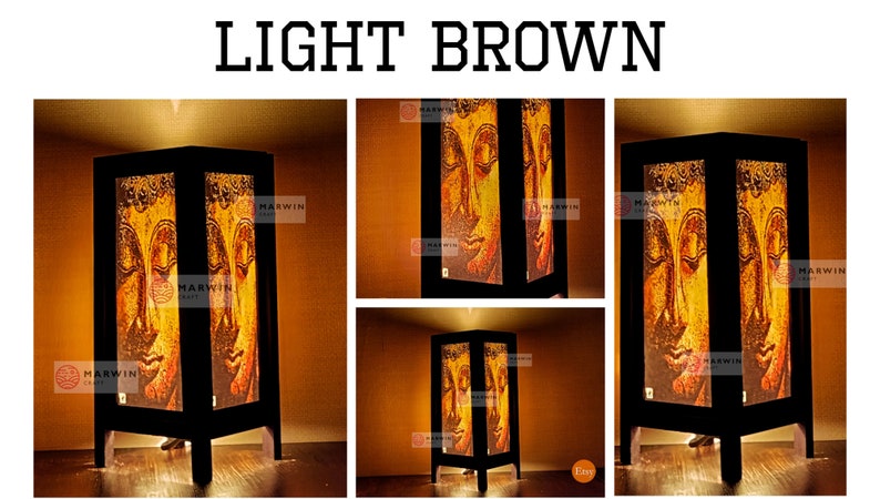 May include: A light brown wooden table lamp with a square base and a rectangular shade. The shade features a printed image of a golden Buddha face. The lamp is lit and casting a warm glow.