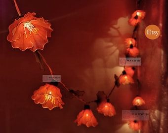 May include: A string of orange flower-shaped fairy lights. Each light has a delicate, layered petal design, with a warm glow emanating from within. The lights are strung together with a dark wire, creating a whimsical and decorative element.