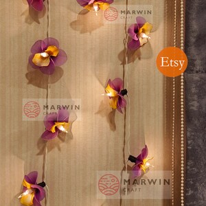 Puede incluir: Una cadena de luces con bombillas en forma de flor de color morado y amarillo. Las luces est&aacute;n enhebradas en un cable blanco y est&aacute;n colgadas en una pared de madera. El texto "MARWIN CRAFT" y "Etsy" es visible en la imagen.