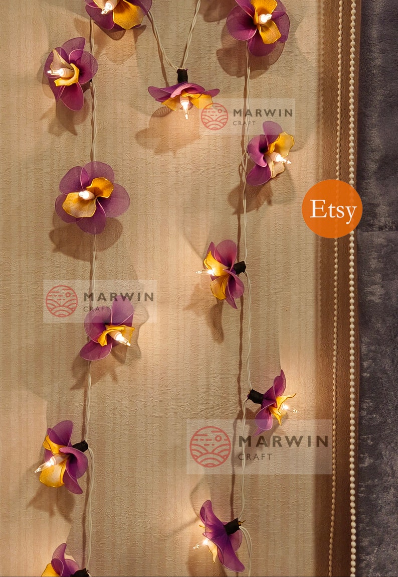 Puede incluir: Una cadena de luces con bombillas en forma de flor de color morado y amarillo. Las luces est&aacute;n enhebradas en un cable blanco y est&aacute;n colgadas en una pared de madera. El texto "MARWIN CRAFT" y "Etsy" es visible en la imagen.