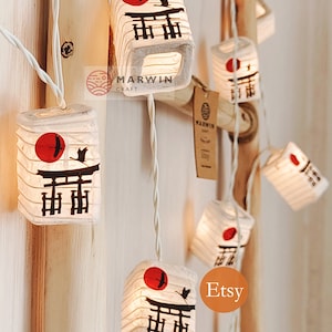 K&ouml;nnte beinhalten: Eine Lichterkette aus wei&szlig;en Papierlaternen mit einer roten Sonne und einer schwarzen Silhouette eines japanischen Tempeltors. Die Laternen sind beleuchtet und h&auml;ngen an einem wei&szlig;en Draht. Der Text "Marwin Craft" und "Etsy" ist auf dem Bild zu sehen.