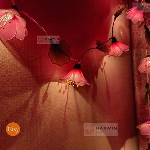 May include: Pink flower string lights with a warm white glow. The lights are strung on a wire and have green leaves between each flower. The flowers are made of fabric and have a delicate, feminine look. The lights are perfect for adding a touch of whimsy to any room.