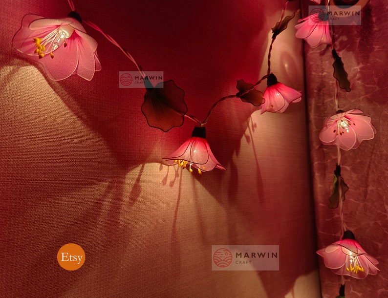 May include: Pink flower string lights with a warm white glow. The lights are strung on a wire and have green leaves between each flower. The flowers are made of fabric and have a delicate, feminine look. The lights are perfect for adding a touch of whimsy to any room.