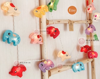 May include: String of lights featuring colorful elephant-shaped paper lanterns. The lights are strung on a white wire and are hanging from a wooden ladder. The text "Etsy" and "Marwin Craft" are visible in the image.