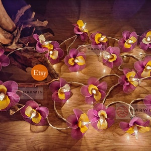 Puede incluir: Una cadena de luces con bombillas en forma de flores moradas y amarillas. Las luces est&aacute;n enhebradas en un cable transparente y est&aacute;n encendidas. Las flores est&aacute;n hechas de tela y tienen un aspecto delicado y femenino. Las luces son perfectas para a&ntilde;adir un toque de fantas&iacute;a y romanticismo a cualquier espacio.