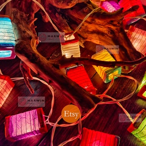 May include: String of colorful paper lanterns with a rustic wooden branch background. The lanterns are lit and glow with warm light. The text "Marwin Craft" and "Etsy" are visible in the image.