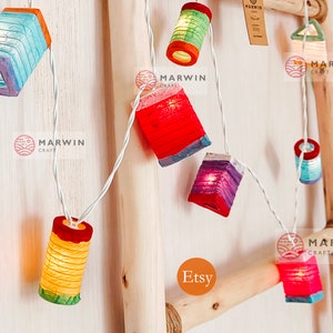 May include: String of colorful paper lanterns in various shapes, including rectangular and cylindrical designs. The lanterns are lit, emitting a warm glow, and strung on a white wire. The background is a light-colored wooden structure.