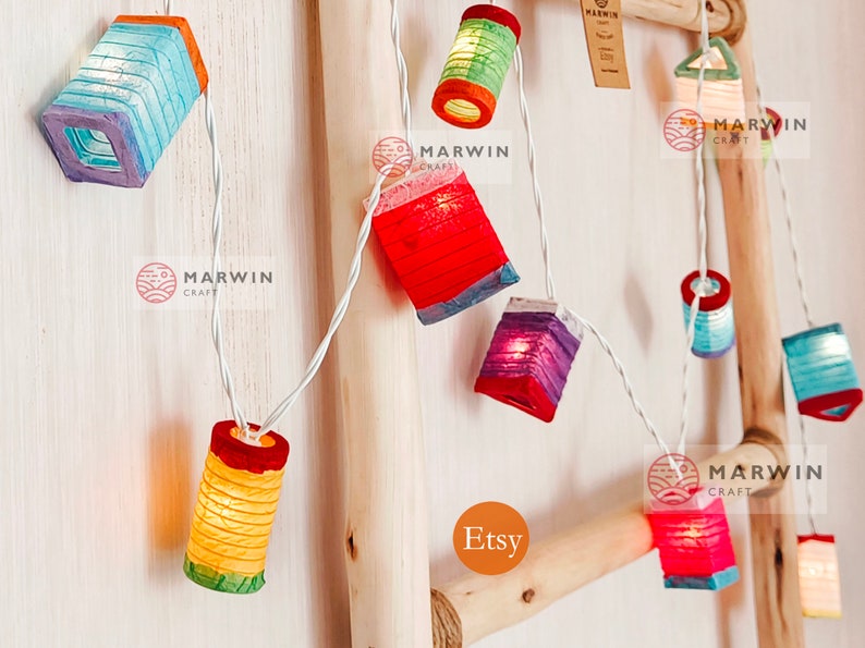 May include: String of colorful paper lanterns in various shapes, including rectangular and cylindrical designs. The lanterns are lit, emitting a warm glow, and strung on a white wire. The background is a light-colored wooden structure.
