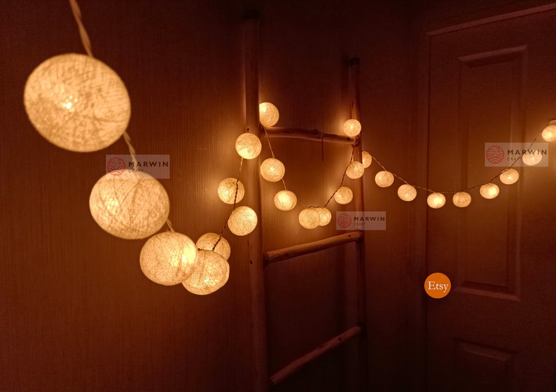 May include: A string of white cotton ball lights with warm white LED bulbs. The lights are strung across a wooden ladder and a doorway. The lights are lit and casting a warm glow.
