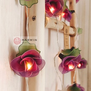 May include: A string of fairy lights with purple and green fabric flowers. The lights are strung on a white wire and are lit. The flowers are arranged in a cascading pattern. The lights are labeled "Marvin Craft" and "Etsy".