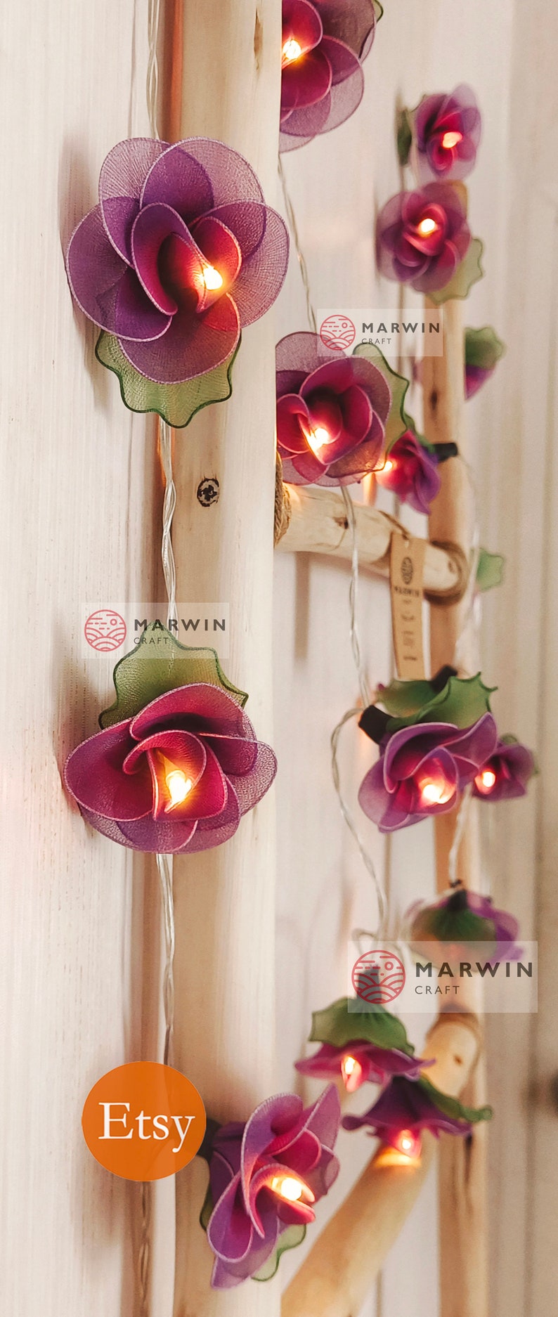 May include: A string of fairy lights with purple and green fabric flowers. The lights are strung on a white wire and are lit. The flowers are arranged in a cascading pattern. The lights are labeled "Marvin Craft" and "Etsy".