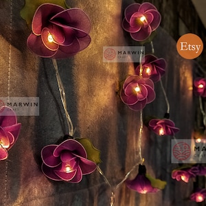 May include: String lights with purple fabric flowers, each with a warm white LED light inside. The lights are strung on a clear wire and are perfect for adding a touch of whimsy to any space. The lights are labeled "Marwin Craft" and "Etsy".