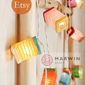 May include: String of colorful, illuminated paper lanterns. Each lantern is a small, square shape with a different color scheme, including red, orange, yellow, and blue. The lanterns are strung on a white wire, with the Etsy logo visible.
