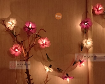 May include: String lights with pink and white flower shaped lights. The lights are strung on a branch and are lit up. The text "Etsy" and "Marwin Craft" are visible in the image.