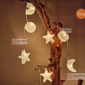 May include: A string of white fabric lights shaped like stars and moons, hanging from a branch. The lights are lit and glowing, creating a warm and inviting atmosphere. The lights are labelled "Marwin Crafts" and "Etsy".