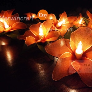 Puede incluir: Flores de papel naranja con luces en su interior, creando un brillo cálido. Las flores están dispuestas en una fila sobre una superficie oscura. El texto "Marwincraft" es visible en las flores.