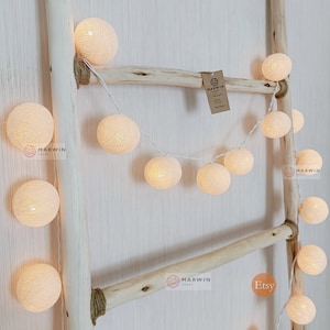 May include: A string of white cotton ball lights draped over a wooden ladder. The lights are battery operated and have a warm white glow. The lights are perfect for adding a touch of whimsy to any room.
