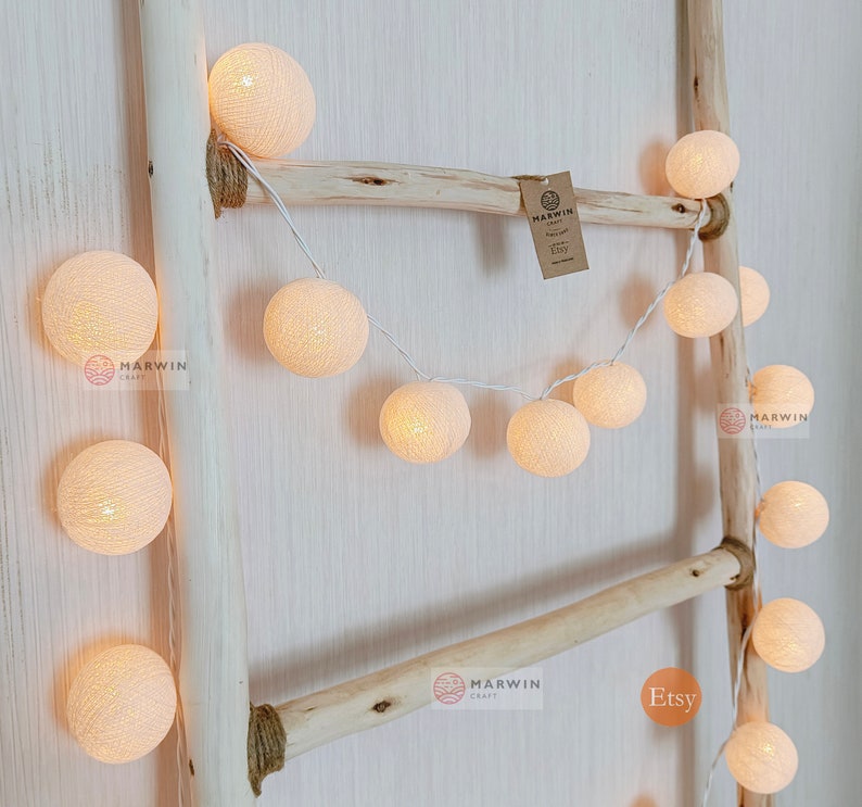 May include: A string of white cotton ball lights draped over a wooden ladder. The lights are battery operated and have a warm white glow. The lights are perfect for adding a touch of whimsy to any room.