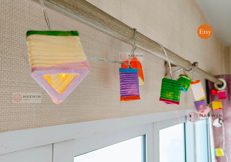 May include: A string of colorful paper lanterns, each with a unique shape and hue, hangs from a rod. The lanterns are lit, casting a warm glow. The Etsy logo is visible in the corner.