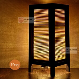 May include: A black wooden table lamp with a rectangular shade made of thin wooden slats. The lamp is lit and casts a warm glow. The lamp is on a wooden surface. The text "Marwin Craft" is visible on the lamp.