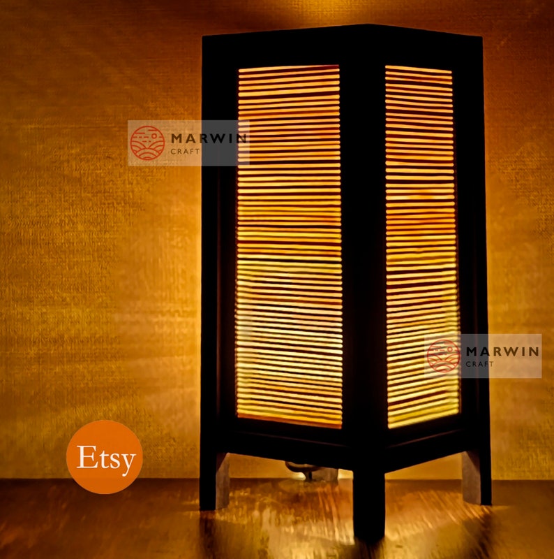 May include: A black wooden table lamp with a rectangular shade made of thin wooden slats. The lamp is lit and casts a warm glow. The lamp is on a wooden surface. The text "Marwin Craft" is visible on the lamp.