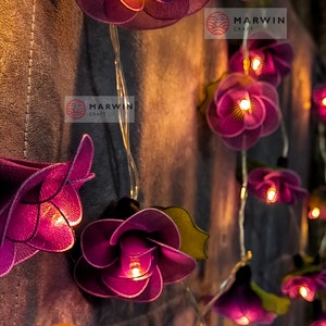 May include: Purple fabric flower string lights with warm white LED lights. The lights are strung on a copper wire and are perfect for adding a touch of whimsy to any space. The flowers are made of a soft, delicate fabric and have a slightly ruffled texture. The lights are battery operated and come with a remote control.