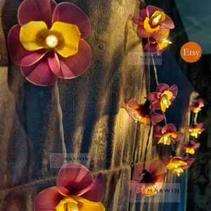 Puede incluir: Una cadena de luces con bombillas en forma de flores moradas y amarillas. Las luces est&aacute;n enhebradas en un cable y est&aacute;n encendidas. Las flores est&aacute;n hechas de tela y tienen un aspecto delicado y femenino. Las luces son perfectas para agregar un toque de fantas&iacute;a y romance a cualquier espacio.