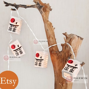 K&ouml;nnte beinhalten: Lichterkette mit wei&szlig;en Lichtern und Papierlaternen mit einem roten Sonnenmotiv, einem schwarzen Vogel und japanischen Schriftzeichen. Die Laternen h&auml;ngen an einem Ast aus Treibholz.