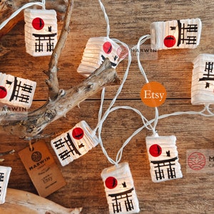K&ouml;nnte beinhalten: Lichterkette mit wei&szlig;en Lichtern und Papierlaternen mit japanischem Design. Jede Laterne hat einen roten Kreis, einen schwarzen Vogel und schwarze Kanji-Zeichen. Die Laternen sind an einem wei&szlig;en Draht befestigt.