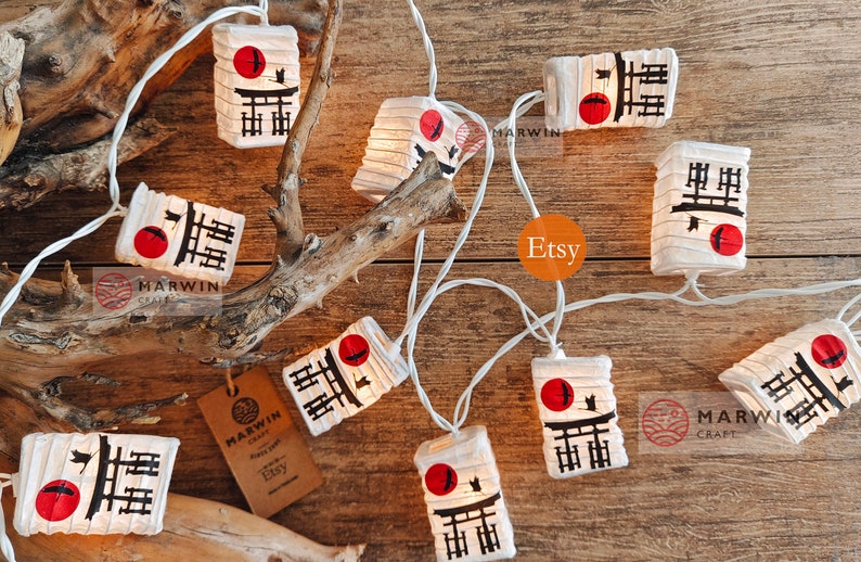 K&ouml;nnte beinhalten: Lichterkette mit wei&szlig;en Lichtern und Papierlaternen mit japanischem Design. Jede Laterne hat einen roten Kreis, einen schwarzen Vogel und schwarze Kanji-Zeichen. Die Laternen sind an einem wei&szlig;en Draht befestigt.
