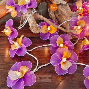 Puede incluir: Una cadena de luces con bombillas en forma de flor de color morado y amarillo. Las luces est&aacute;n enhebradas en un cable blanco y est&aacute;n encendidas. Las flores est&aacute;n hechas de tela y tienen un aspecto delicado y femenino. Las luces son perfectas para agregar un toque de fantas&iacute;a y encanto a cualquier espacio.
