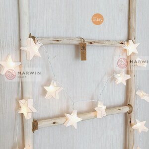 May include: White string lights with star-shaped paper lanterns. The lights are strung on a wooden ladder and the word "Etsy" is written in a circle above the lights.