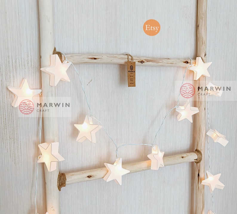 May include: White string lights with star-shaped paper lanterns. The lights are strung on a wooden ladder and the word "Etsy" is written in a circle above the lights.
