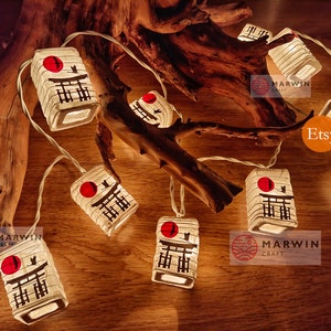 K&ouml;nnte beinhalten: Lichterkette aus wei&szlig;en Papierlaternen mit einer roten Sonne und schwarzen japanischen Schriftzeichen. Die Laternen sind beleuchtet und h&auml;ngen an einem Ast. Der Text "Marwin Craft" und "Etsy" ist im Bild sichtbar.